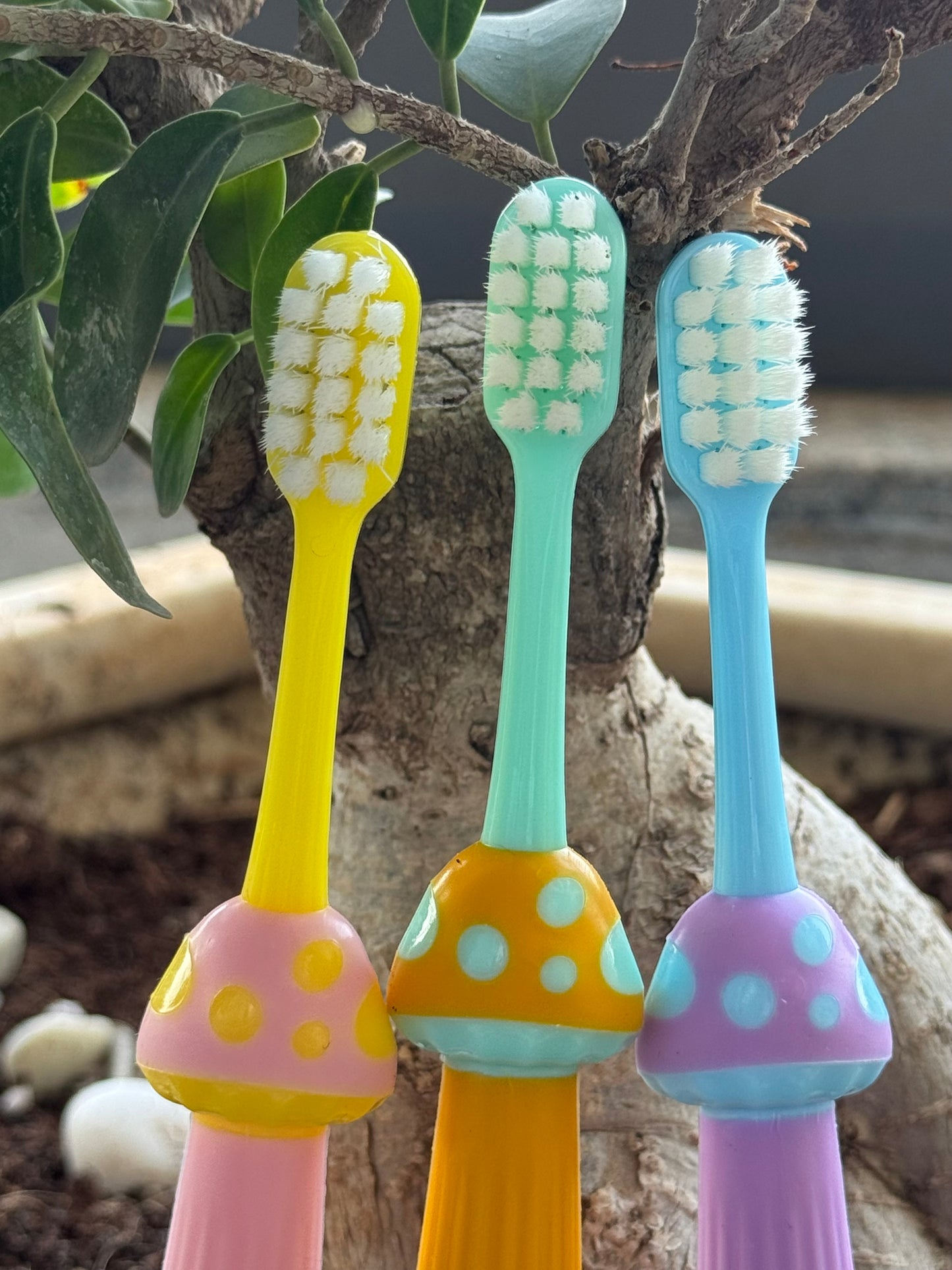 Three colorful children's toothbrushes with mushroom-shaped handles against a natural background.